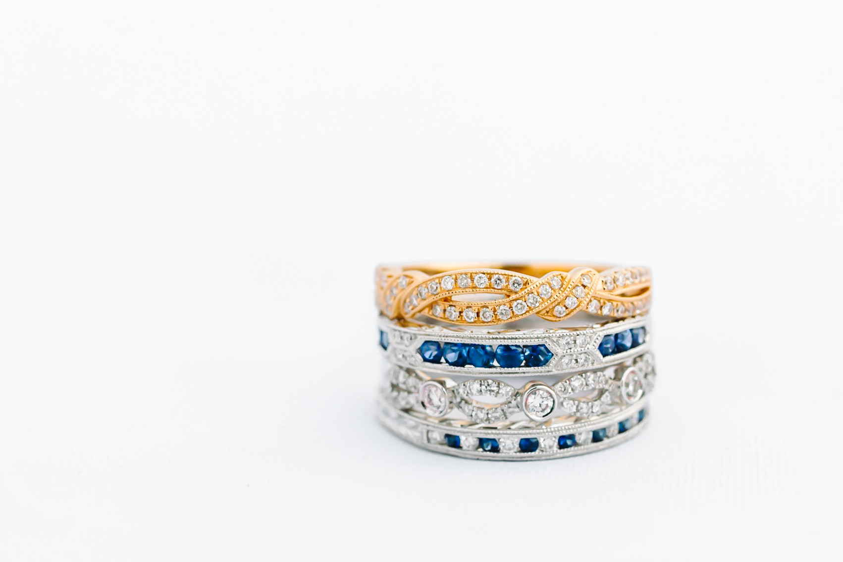 Stack of Kirk Kara yellow and white gold wedding bands, some with sapphires, on a white background